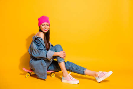 Full length side profile body size photo beautiful she her lady weekend vacation sporty free time sitting own skate board wear casual jeans denim jacket shoes pink hat isolated yellow vivid backgroundの写真素材