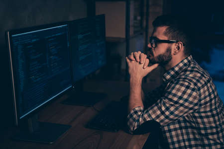 Close up side profile photo handsome he him his guy ponder pensive hands arms chin coder code development outsource IT processing two monitors table office wear specs formalwear plaid shirtの写真素材