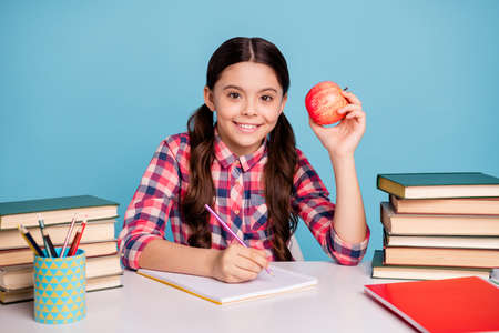 Portrait of nice attractive lovely winsome cheerful cheery smart clever girl wearing checked shirt doing home work eating ripe apple isolated over bright vivid shine blue turquoise backgroundの写真素材