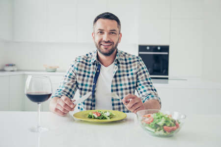 Portrait of charming imposing mature guy sit table hold hand fork knife want cut pleasant evening cheerful checked shirt big light apartment wineglassの写真素材