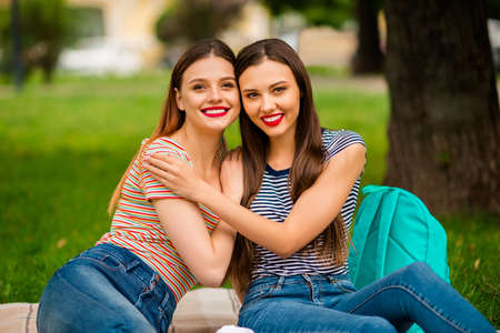 Portrait of sweet girls with red lips-stick cuddling wearing striped t-shirt denim jeans sitting on plaid blanket in townの写真素材