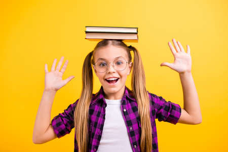 Closeup photo of little lady pupil hold book on head laughing raise arms air wear specs checkered shirt isolated yellow backgroundの写真素材