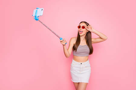 Portrait of funny student holding monopod with device making photo grimacing wearing red eyewear eyeglasses isolated over pink backgroundの写真素材