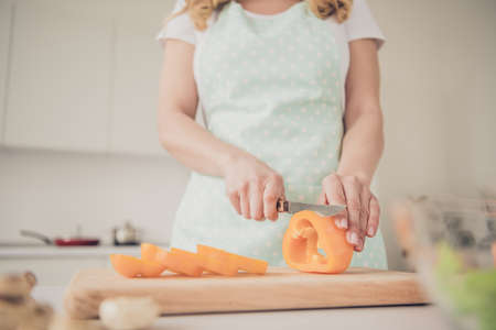 Cropped close-up view of her she cutting making homemade useful edible farm products orange juicy pepper on board in light white interior kitchen indoorsの写真素材
