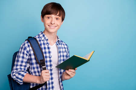 Portrait of pretty boy holding printed book smiling wearing checked shirt isolated over blue...
