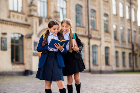 Portrait of pretty kids with blonde brunette pigtails ponytails hold backpack rucksack read books wear black blazers jackets skirt knee-socks outdoorsの写真素材