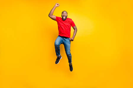 Full length body size photo of jumping man wearing red t-shirt jeans denim rejoicing with his victory at something while isolated with yellow background