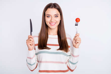 Portrait of charming woman holding tomato looking at camera with toothy smile wearing striped jumper...