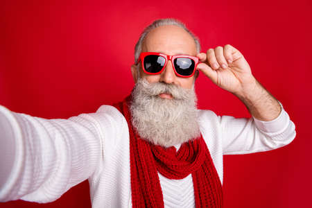 Fashion aged santa man making self photos wear sun specs knitted jumper isolated red backgroundの写真素材