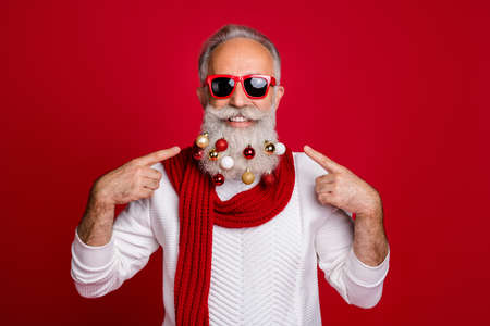 Portrait of attractive retired man in eyeglasses eyewear pointing at his beard with collection of glass balls wearing white pullover isolated over red backgroundの写真素材