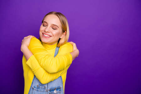 Close up photo of cheerful lady cuddle herself with closed eyes wear yellow turtleneck denim jeans overalls isolated over purple violet backgroundの写真素材