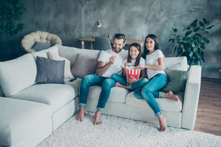 Portrait of nice attractive lovely cheerful family mommy daddy wearing casual white t-shirts jeans denim sitting on sofa enjoying video spending free time weekend at industrial style interior roomの写真素材