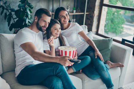Portrait of nice attractive lovely charming cheerful cheery family wearing casual white t-shirts jeans denim sitting on divan having fun watching series serial video switching channel indoorsの写真素材