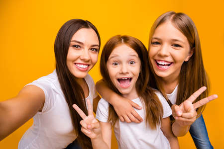 Photo of three sister ladies, making selfies showing v-sign symbol wear casual white t-shirts isolated yellow backgroundの写真素材
