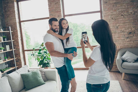 Portrait of nice attractive cheerful cheery friendly family in casual white t-shirts jeans brunette mommy taking making capture at industrial loft style interior house living-room indoorsの写真素材