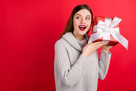 Pretty lady with big giftbox wondering what inside wear knitted sweater isolated red background