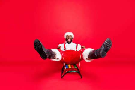 Full length photo of african mixed race santa in nicholas cap screaming sledding wearing pants trousers white sweater isolated over red backgroundの写真素材