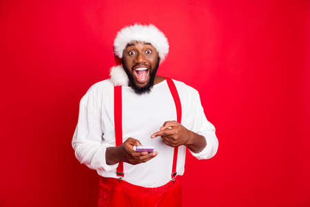 Portrait of impressed black santa claus in xmas hat pointing at his cell phone device screaming wow...