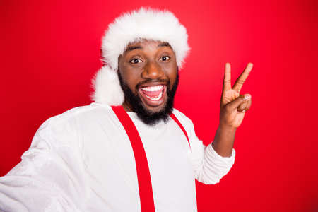 Close up photo of excited black christmas father in xmas hat enjoying memory take selfie making v-signs wearing white jumper isolated over red backgroundの写真素材