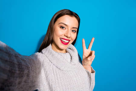 Photo of pretty lady making selfies showing v-sign symbol wear warm knitted pullover isolated blue backgroundの写真素材