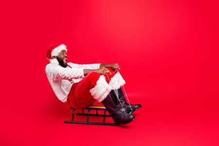 Full length photo of excited black santa claus in xmas cap screaming wow riding sledge wearing white pullover pants trousers isolated over red backgroundの写真素材