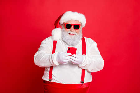 Portrait of funny funky fat overweight santa claus with big abdomen belly use cell phone comment newyear posts wearing stylish trendy eyewear eyeglasses hat reading news isolated over red backgroundの写真素材