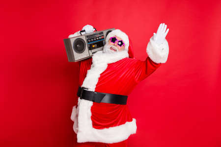 Forever young. Portrait of crazy style santa claus in eyewear eyeglasses raising palms holding retro cassette dancing having christmas party isolated over red backgroundの写真素材