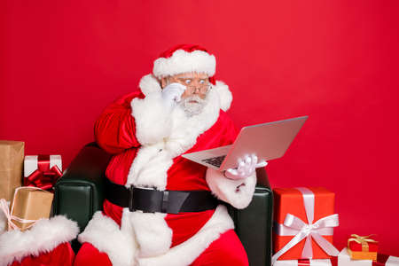 Portrait of astonished santa claus touching his eyewear eyeglasses looking news sales discounts at netbook wonder wearing hat belt isolated over red backgroundの写真素材
