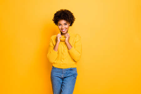Photo of dark skin funny lady in good mood holding hands big collar of warm knitted pullover isolated yellow color backgroundの写真素材