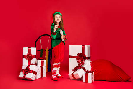 Full length body size view of nice attractive sweet curious funny small little pre-teen elf writing creating winter idea wish list pile stack purchase isolated over bright vivid shine red backgroundの写真素材