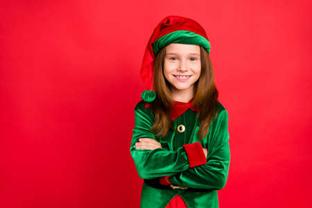 Portrait of cheerful elf girl in hat with her hands crossed smiling ready to help in christmas night wearing cap costume isolated over red backgroundの写真素材