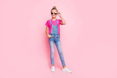 Full length body size photo of cheerful cute funny girl standing confidently in front of camera with hand in pocket wearing jeans overall footwear holding eyewear isolated over pastel pink color backgroundの写真素材