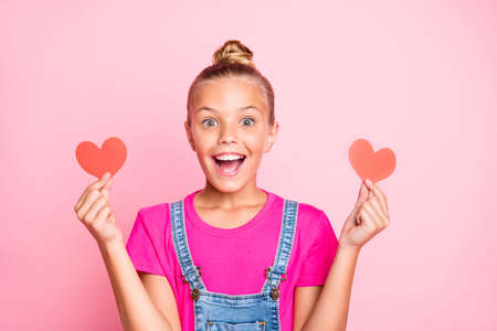 Photo of excited cheerful cute nice schoolgirl holding two hear shapes red with her hands wearing jeans denim overall fuchsia t-shirt isolated over pink pastel color backgroundの写真素材