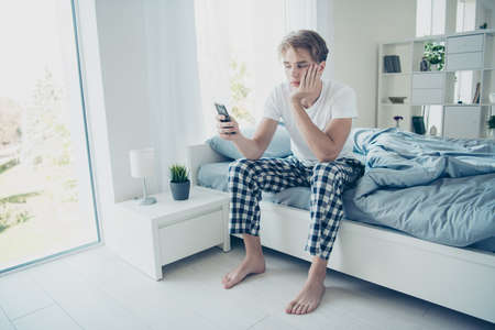 Full length photo of tired unhappy upset blonde haired man have night work try not sleep use his cellphone follow social network accounts blogs sit on bed in house indoorsの写真素材