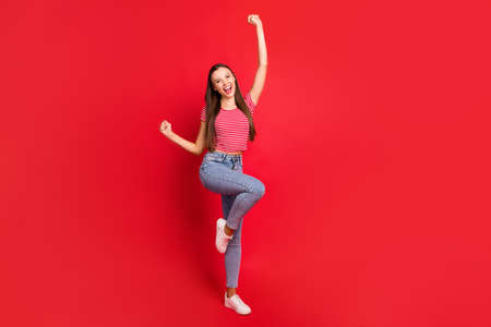 Full length body size photo of rejoicing trendy overjoyed charming beautiful girl dancing with success wearing striped t-shirt while isolated with red backgroundの写真素材