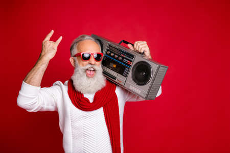 Portrait od foolish santa in eyewear eyeglasses holding boombox showing heavy metal sign wearing white pullover isolated over red backgroundの写真素材