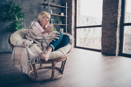 Full length profile side photo of charming peaceful dreamy middle aged woman sit in armchair covered with checkered blanket hold mug look window dream imagine autumn holidays indoorsの写真素材