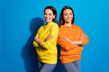 Photo of pretty two girlfriends lady standing back-to-back in good mood, crossing arms wear casual bright hoodies and jeans isolated blue color backgroundの写真素材