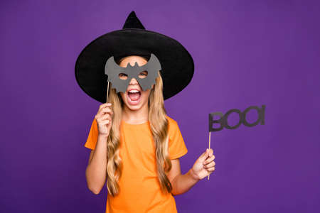 Photo of little witch lady play paranormal role halloween theme party, holding bat paper stick scary look wear orange t-shirt wizard hat isolated purple color backgroundの写真素材