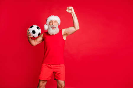 Portrait of crazy fan santa claus in hat cap screaming yes raising fists holding, ball see teams goal wearing sportswear isolated over red backgroundの写真素材