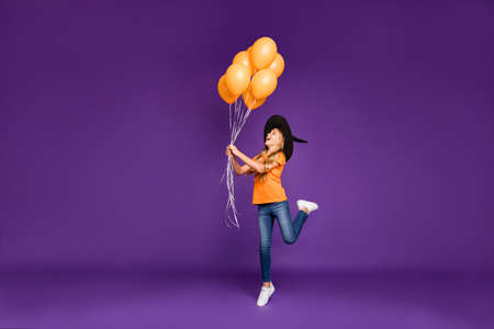 Full body photo of little witch lady halloween party holding air balloons, making spell to make them fly wear orange t-shirt wizard hat isolated purple color backgroundの写真素材