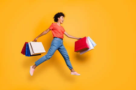 Full length profile side photo of amazed funky funny wavy brunette hair girl hold bags jump run to shopping center boutique, hurry wear red t-shirt denim jeans isolated over yellow color backgroundの写真素材
