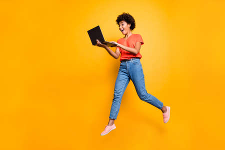 Full length photo of excited funny funky curly hair girl use her laptop find news information about sales jump wear denim jeans stylish, trendy sneakers t-shirt isolated over yellow color backgroundの写真素材