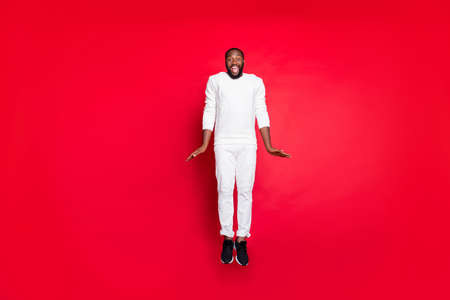 Full size photo of excited dark skin handsome guy jumping high rejoicing having best x-mas party time wear white sweater trousers isolated red backgroundの写真素材