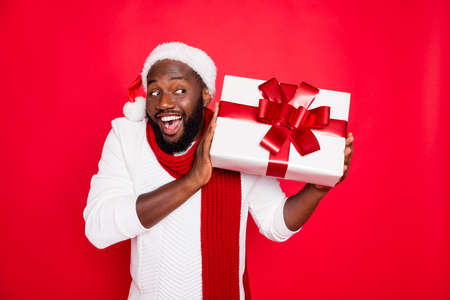 Portrait of amazed funky dark skin man wear santa claus headwear hold big package receive on x-mas time celebration scream wow feel interested wear style pullover isolated over red color backgroundの写真素材