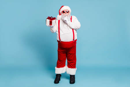 Full length photo of funny funky fat modern santa claus with big belly show index finger, speechless sign hold gift box want give wish for eve wear red cap suspenders isolated blue color backgroundの写真素材