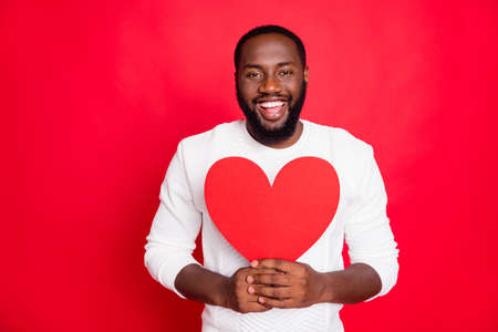 Portrait of positive candid mulatto man hold big paper card heart present for his girlfriend on 14-february wear white outfit isolated over red color backgroundの写真素材