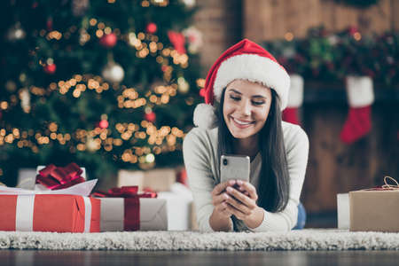Close up photo of positive girl in santa claus hat lie on carpet use smartphone send christmas...