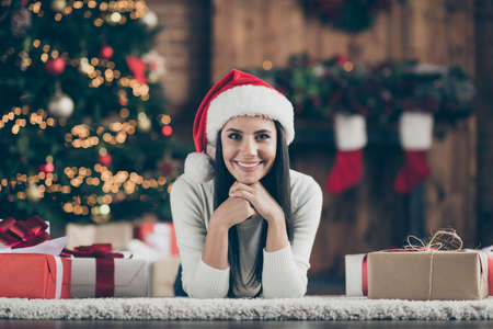 Close up photo of positive cheerful girl lie on floor carpet with cristmas gift boxes enjoying x-mas time jolly party wear white pullover in house with newyear garlands illumination indoorsの写真素材