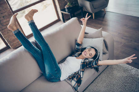 High angle view photo of amazing lady listening favorite radio fm in modern earflaps raising legs up showing v-signs lying comfy sofa wear casual clothes apartment indoorsの写真素材